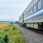 A modern Canadian passenger train traveling through scenic prairie landscape with hemp plants visible near the route in soft focus.